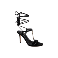 The Bond Suede Sandal is a black high-heeled shoe with a strappy design, featuring an open toe, stiletto heel, gold chain detail on the front, and a thin ankle wrap strap that ties around the ankle with tassel accents.