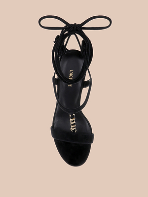 Bond Suede Sandal with high heel, ankle wrap straps, and chain detail on the insole, shown from above on a light beige background. Elegant straps create a striking silhouette.