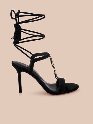 The Bond Suede Sandal in black features a pointed strappy heel, ankle wrap-around straps, gold chain detailing at the front, and chic tassel accents—set against a plain beige background.