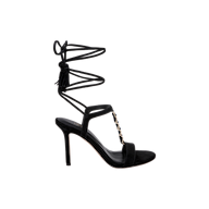 The Bond Suede Sandal is a black high-heeled shoe featuring a thin ankle wrap strap, gold chain detail across the front, an open toe, and stiletto strappy heel, displayed against a plain white background.