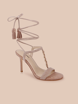 La sandale Bond Suede en rose blush est dotée de brides enveloppantes au niveau de la cheville, dextrémités à pompon et dune chaîne dorée sur le devant - un talon à lanières très tendance, présenté sur un fond pêche clair.