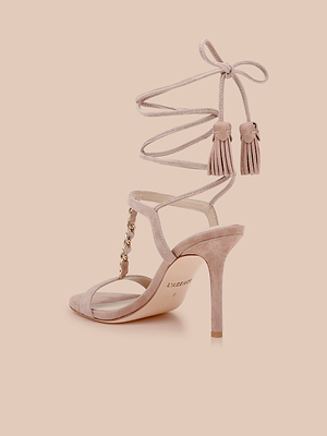 La sandale Bond Suede en rose blush présente un talon à lanières, des lanières enveloppantes à la cheville avec des accents de houppes, et des détails de chaînes dorées sur la tige, le tout sur un fond beige clair.