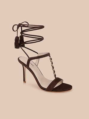 The Bond Suede Sandal showcases a suede upper, ankle wrap with tassel ends, thin front strap, gold-detailed T-strap, and open toe—all on a chic strappy heel and light beige background.