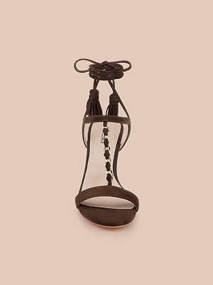 Front view of the Bond Suede Sandal with a suede upper, ankle lace-up strap, decorative tassels, and chain detail on the strappy heel, set against a solid light beige background.