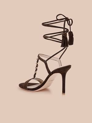 The Bond Suede Sandal features a brown suede upper, open toe, gold studded strap, and long ankle ties with tassels, shown on a light beige background.
