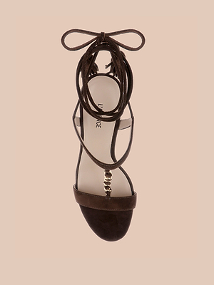 The Bond Suede Sandal is displayed from above on a beige background, featuring a suede upper, flat sole, ankle wrap laces, and metallic bead accents.