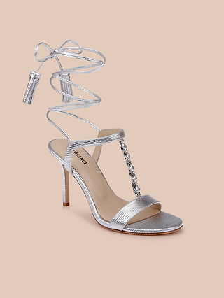 The Bond Leather Sandal is a silver leather sandal with an elegant strappy heel design, ankle-wrapping lace, a chic chain detail on the front strap, and a sleek stiletto heel, beautifully displayed against a beige background.