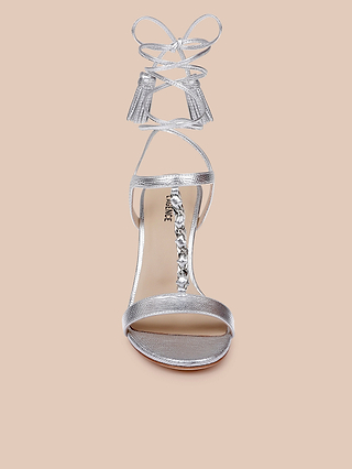 The Bond Leather Sandal is a silver strappy heel crafted from leather, designed with ankle wrap laces and a decorative jewel chain embellishing the T-strap front, showcased against a light beige background.