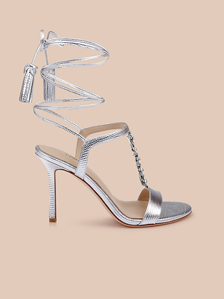 Introducing the Bond Leather Sandal: a silver leather high-heeled sandal featuring an ankle wrap strap, open toe, eye-catching chain detail on the T-strap, and elegant tassel accents at the ends of the wrap. This strappy Bond Leather Sandal is beautifully displayed on a beige background.