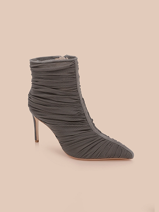 The Fulton Mesh Bootie is a black stiletto bootie with a high heel, featuring a ruched, textured mesh upper and a pointed toe for added sophistication. It is displayed against a plain light beige background.