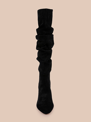 The Fletcher Boot, featuring a slouchy, knee-high silhouette in black suede, is displayed upright against a plain beige background and viewed from the front. This block heel design highlights its soft suede material and effortlessly chic appearance.