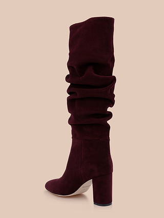 The Fletcher Boot is a tall, slouchy suede boot in rich maroon with a bold high block heel, embodying bohemian chic style and shown against a plain beige background.