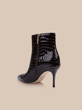 The Houston Bootie, crafted from glossy black patent leather with a crocodile texture, features a pointed toe, mid-height stiletto heel, and a side zipper. It is displayed against a beige background.