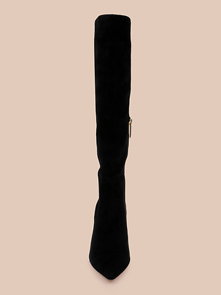 The Herald Boot, a single knee-high boot in black suede with a pointed toe and sleek stiletto heel, is displayed upright against a light beige background. A side zipper is visible on the boot.