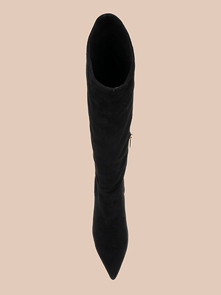 The Herald Boot, a black suede knee-high style featuring a pointed toe and sleek stiletto heel, is displayed from a top-down perspective against a beige background.