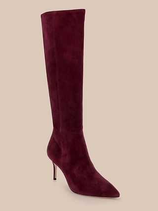The Herald Boot, a knee-high Burgundy suede style featuring a pointed toe and elegant stiletto heel, is displayed against a plain beige background.