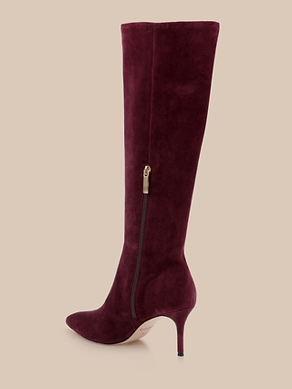 The Herald Boot, a single knee-high suede boot in dark burgundy with a pointed toe stiletto and side zipper, is displayed on a beige background.