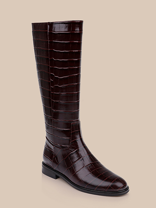 The Cortland Boot is a knee-high riding boot crafted from dark brown croc-embossed leather with a glossy finish and a low heel, displayed against a plain beige background.