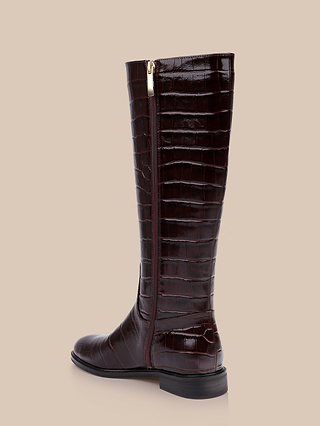 The Cortland Boot is a tall, dark brown, knee-high boot made of crocodile-embossed leather, featuring a gold side zipper, low heel, and slightly pointed toe, displayed against a beige background.