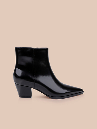 The Vesey Bootie—a shiny black leather ankle boot with a pointed toe and a low, chunky heel—is displayed on a plain beige background, embodying the timeless style of classic western ankle boots.