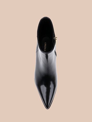 A top view of the Vesey Bootie—a shiny black leather ankle boot with a pointed toe, side zipper, and gold buckle detail—displayed on a beige background.
