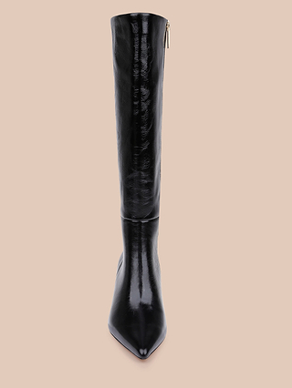 The Baxter Boot, a black, knee-high, pointed-toe patent leather style, is displayed upright against a solid beige background in a front-facing photograph.