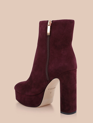 The Soho Bootie is a burgundy suede platform ankle boot with a chunky block heel, gold side zipper, and beige sole, showcased against a beige background.