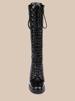 Front view of the Tribeca Boot—a tall, black, quilted lace-up combat boot featuring a shiny patent leather finish and rugged sole—displayed against a plain beige background.