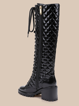 The Tribeca Boot—a shiny black patent leather knee-high boot featuring a quilted pattern, chunky block heel, side zipper, and lace-up front—is displayed on a neutral beige background.
