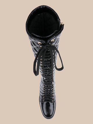 The Tribeca Boot, a black knee-high combat style with a quilted texture, patent leather lace-up front, and glossy toe cap, is shown from above against a beige background.