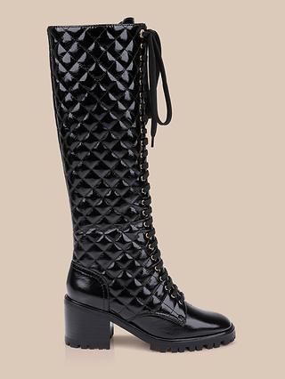 The Tribeca Boot is a tall, black, quilted combat boot featuring a chunky block heel and front laces, expertly crafted from sleek patent leather and displayed on a plain beige background.
