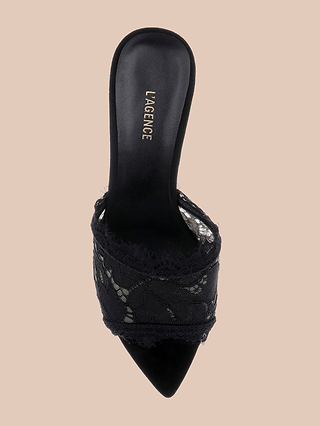 The Penn Mule is a black lace mule with a pointed toe and elegant stiletto heel, featuring delicate lace detailing and the LAGENCE brand name printed on the insole, shown against a beige background.