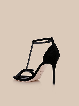 The Lerner Sandal is a black suede high-heeled T-strap sandal with an ankle strap and open toe, featuring a sleek stiletto heel and a soft suede upper, displayed on a neutral beige background.