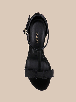 The Lerner Sandal is a black open-toe style featuring a suede upper, ankle strap, and bow detail on the toe strap. The insole displays the LAGENCE logo and the sandal is shown from above against a beige background.
