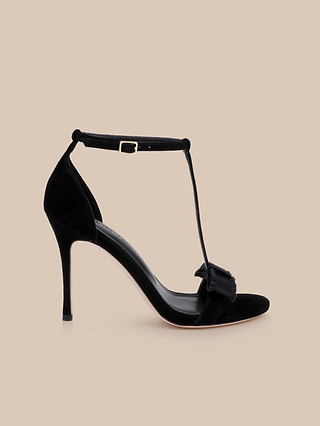 The Lerner Sandal is a black high-heeled sandal with a sleek stiletto heel, showcasing an ankle strap, T-strap design, and a decorative bow on the front. This elegant T-strap sandal is displayed against a plain beige background.