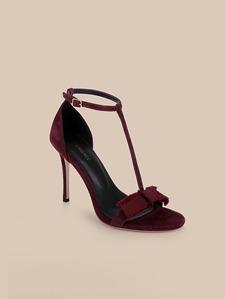 The Lerner Sandal showcases a burgundy suede upper with a classic T-strap design, ankle buckle closure, and a decorative bow adorning the toe. This elegant high-heeled stiletto is displayed against a plain beige background.