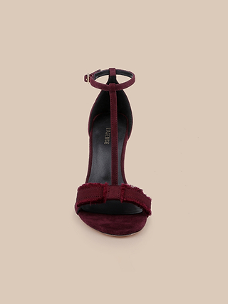 Front view of the Lerner Sandal in burgundy, featuring a T-strap design with an ankle strap, stiletto heel, frayed bow detail over the toe, and a soft suede upper, displayed against a plain beige background.