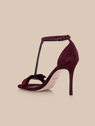 The Lerner Sandal is a burgundy T-strap sandal with a stiletto heel, crafted from soft suede, featuring an ankle strap with buckle and an open toe, displayed against a beige background.
