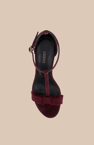 The Lerner Sandal, a single womens stiletto heel in burgundy suede with an ankle strap and bow detail, is displayed from above on a beige background. The black footbed features LAURENCE printed in gold lettering.
