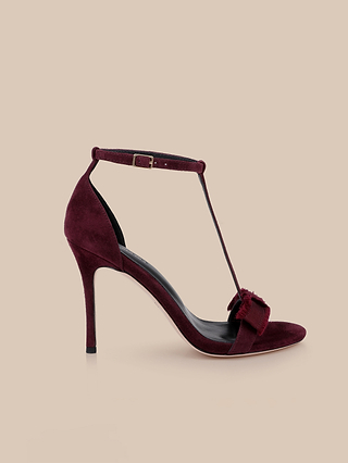 The Lerner Sandal features a burgundy suede upper with a high heel, T-strap design, ankle buckle, and a decorative bow on the toe, displayed against a beige background.