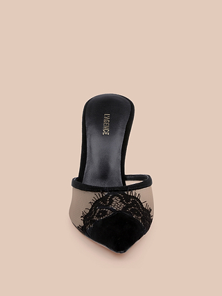 Displayed on a beige background is the Semois Mule—a black suede heel mule featuring a pointed toe, mesh side panels, and refined lace detailing across the front. This stiletto-heeled lace-capped mule seamlessly combines sophistication with modern style.
