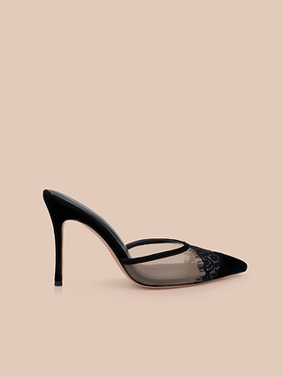 The Semois Mule features black suede with a pointed toe, mesh side panels, intricate lace detailing, and a bold stiletto heel, all beautifully showcased against a beige background.