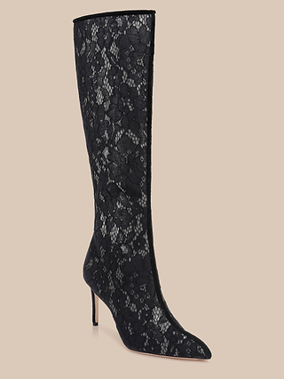 The Chambers Boot, a black knee-high high-heeled boot featuring a pointed toe and adorned with an intricate lace pattern, is displayed against a light beige background.