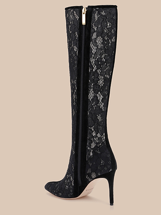 The Chambers Boot is a striking black lace boot with a pointed toe and stiletto heel, featuring a delicate lace overlay and a side zipper with a gold pull. This elegant high-heeled boot is beautifully displayed against a beige background.