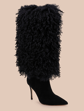 The Sheridan Boot features a sleek black high heel, a pointed toe, and a tall shaft wrapped in shaggy, curly-textured faux fur, showcased against a plain beige background.