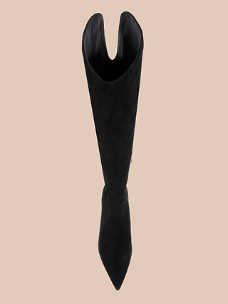 The Grand Boot is a black knee-high suede boot with a pointed toe and low heel, viewed from above against a beige background. Its top opening forms a distinctive V-shape.