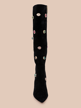 The Concourse Boot is a black knee-high boot featuring vibrant jeweled embellishments and a distinctive eye-shaped detail, set against a beige backdrop—these luxury statement boots elevate style with their captivating charm.