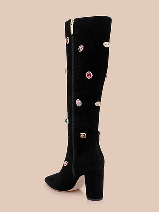 The Concourse Boot, a black knee-high suede boot featuring a chunky heel, colorful gem-like stud embellishments, and a gold zipper adorning the upper side, is showcased against a beige background.