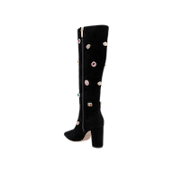 The Concourse Boot is a black knee-high boot with a chunky heel, adorned with vibrant gem-like embellishments and a gold zipper running down the side. Presented against a plain white background, these luxury statement boots are designed to elevate any outfit with their bold, sparkling accents.
