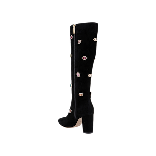 The Concourse Boot is a black suede knee-high boot with a chunky high heel, adorned with colorful crystal-inspired embellishments and equipped with a convenient side zipper, displayed against a plain white background.